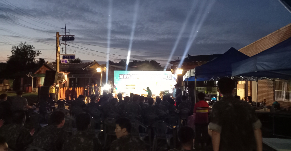 제3회 어은마을 한마음 축제 공연이 밤늦게 공연이 진행되고 있고 군인들과 마을사람들이 좌석에 앉아 공연을 관람하고있는 모습